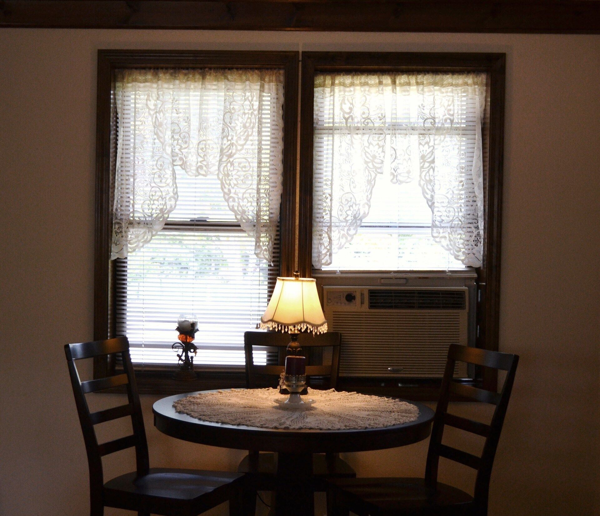 A room with a round table, two chairs, a table lamp, and a window with decorative curtains.