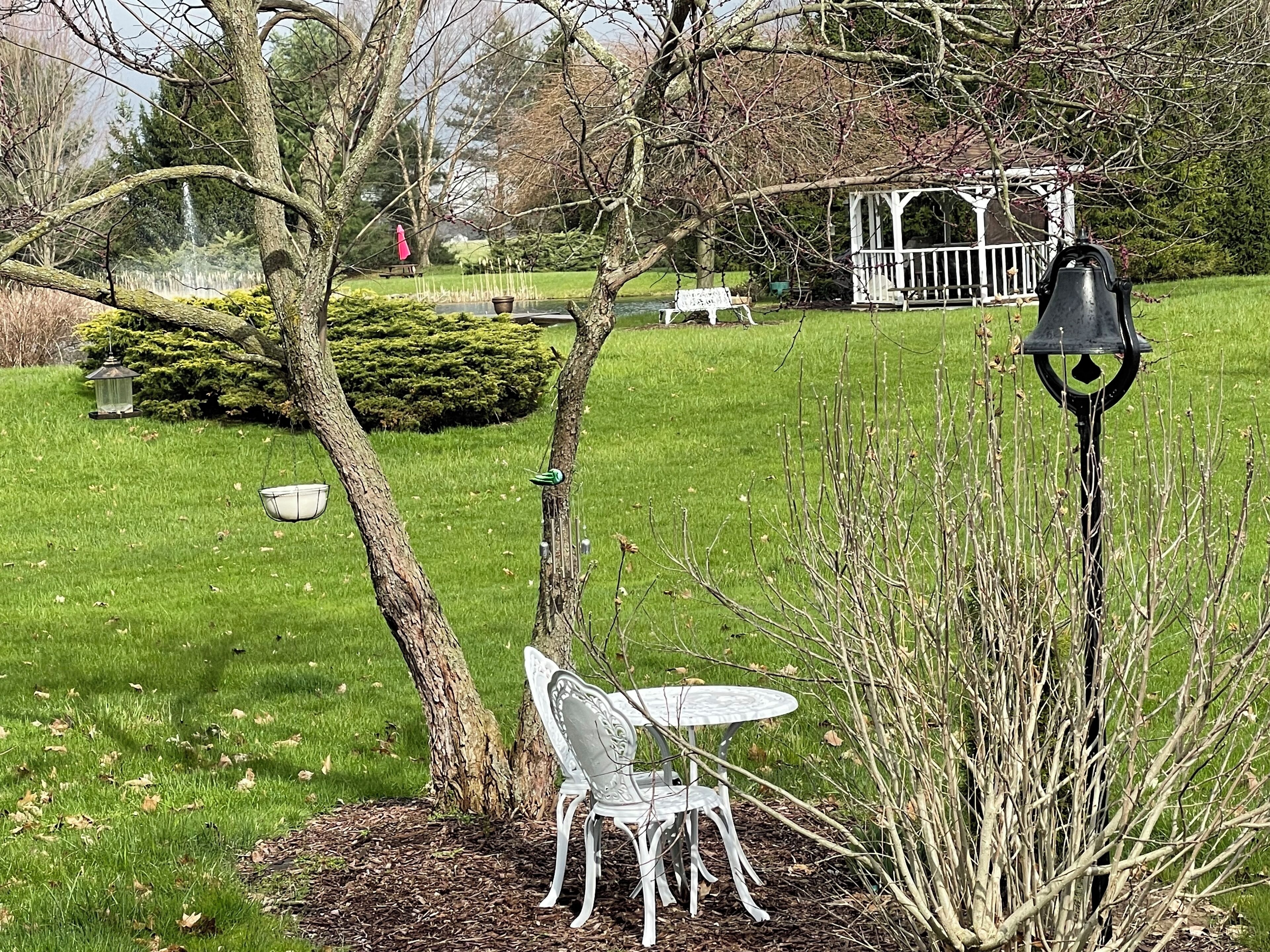 A garden with a white metal table and chair, a bird feeder, and a gazebo in the background.