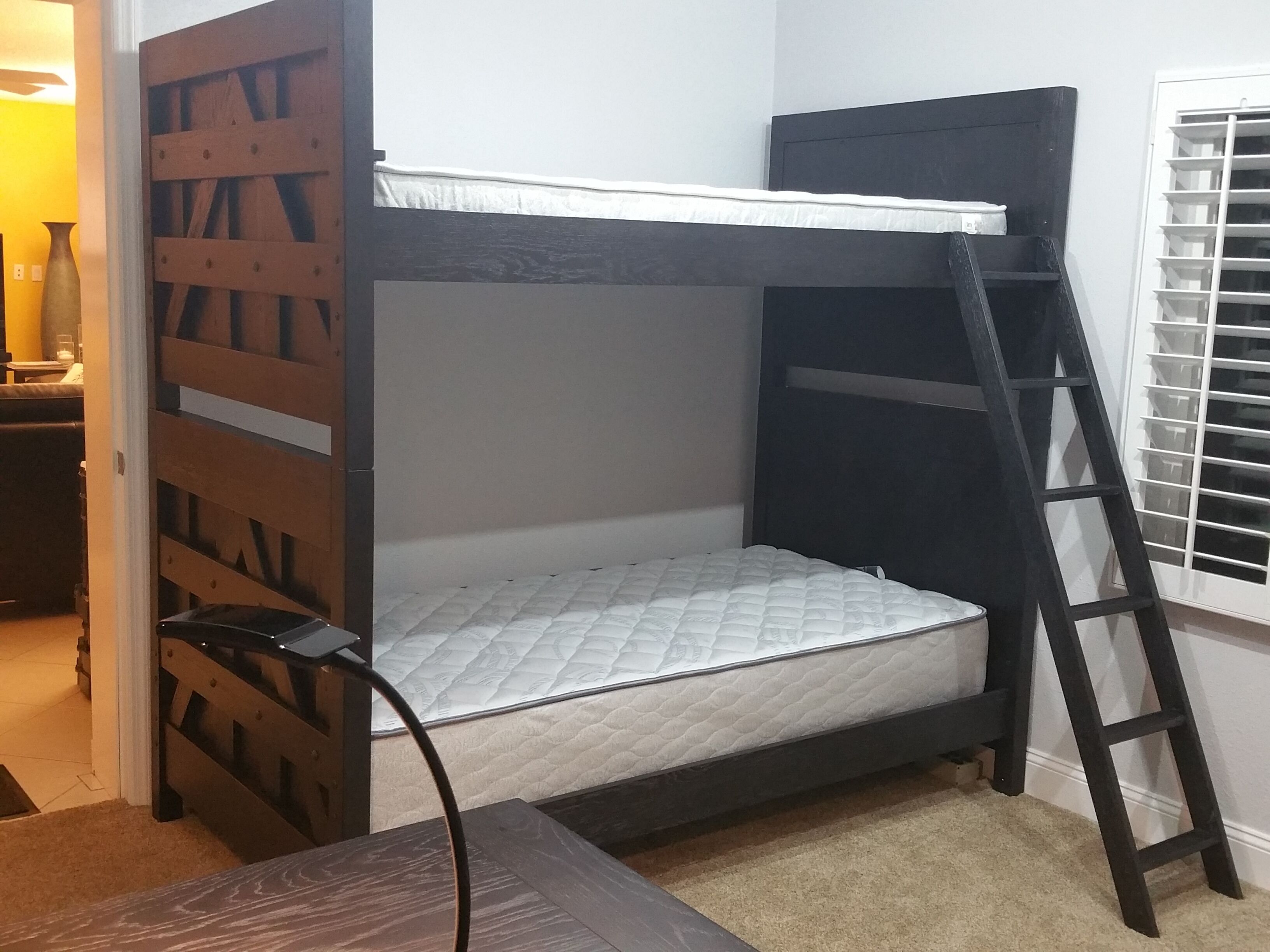 A bunk bed with a ladder, a bedside table, and a window with blinds.