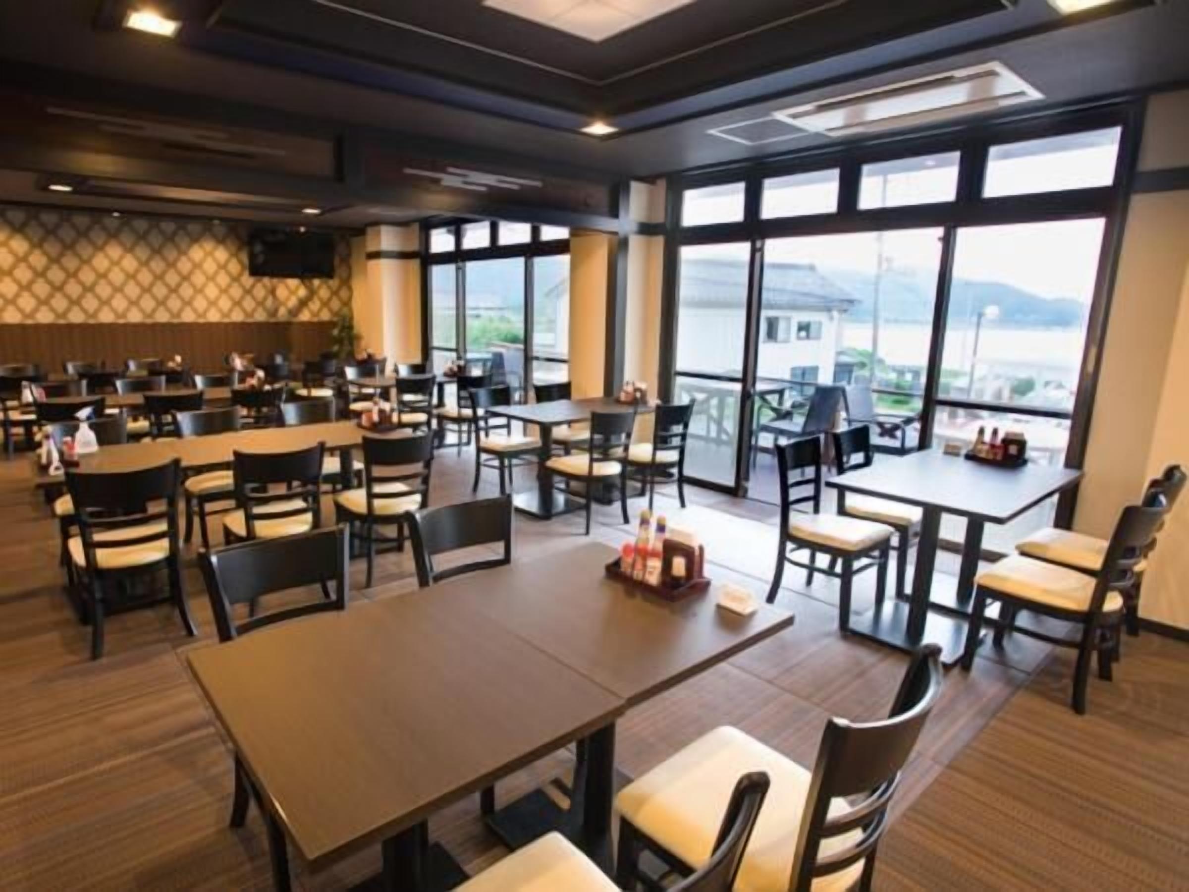 A spacious dining area with multiple tables and chairs, large windows offering a view of the outdoors, and a modern ceiling design.