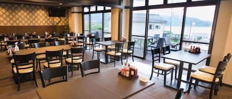 A spacious dining area with multiple tables and chairs, large windows offering a view of the outdoors, and a modern ceiling design.