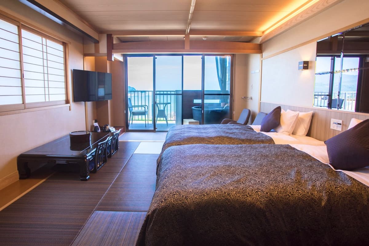 A hotel room with two beds, a desk with a chair, a television, and a balcony with a view.