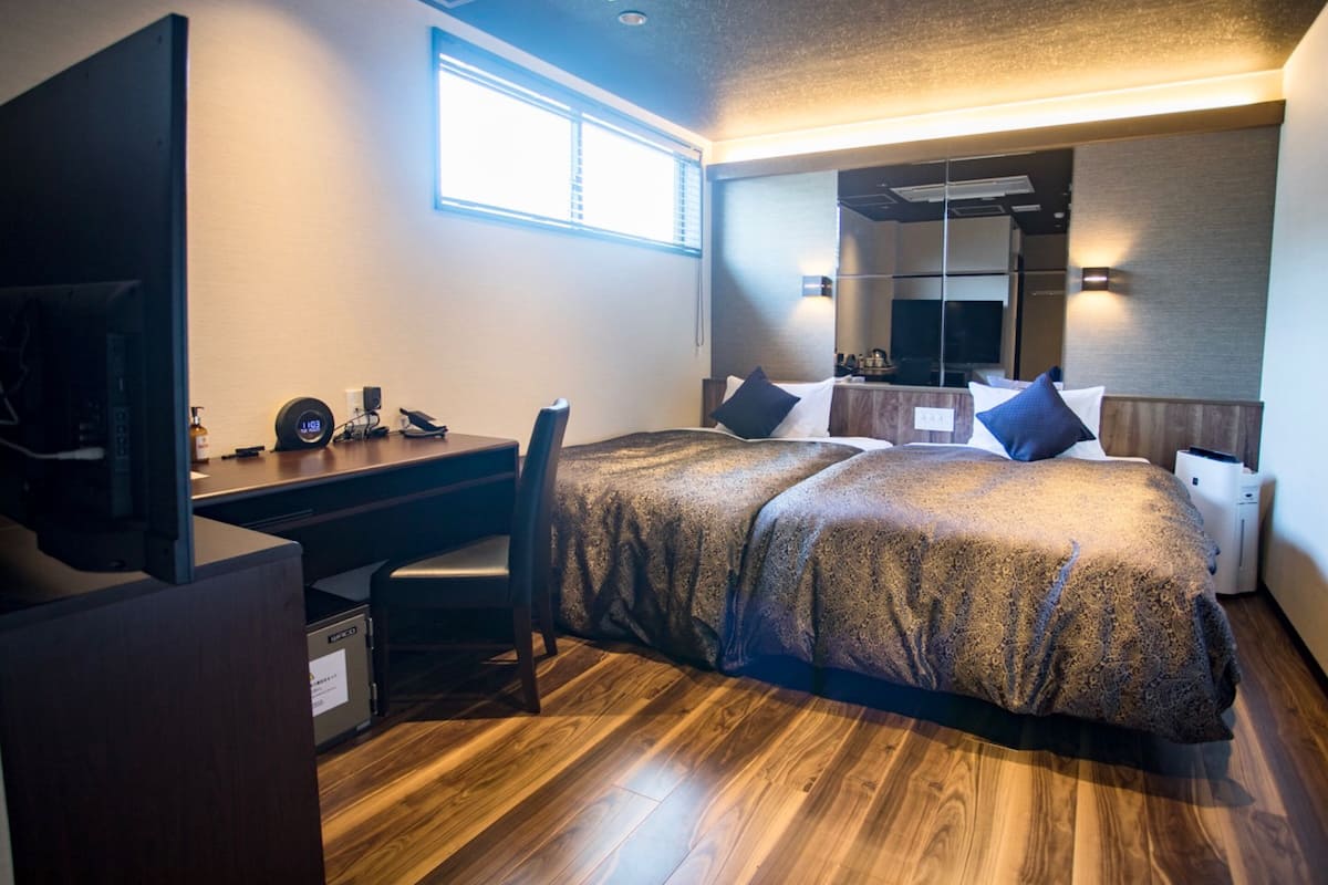 A modern hotel room with a wooden floor, a large bed with patterned bedding, a desk with a computer and phone, and a large mirror above the bed.