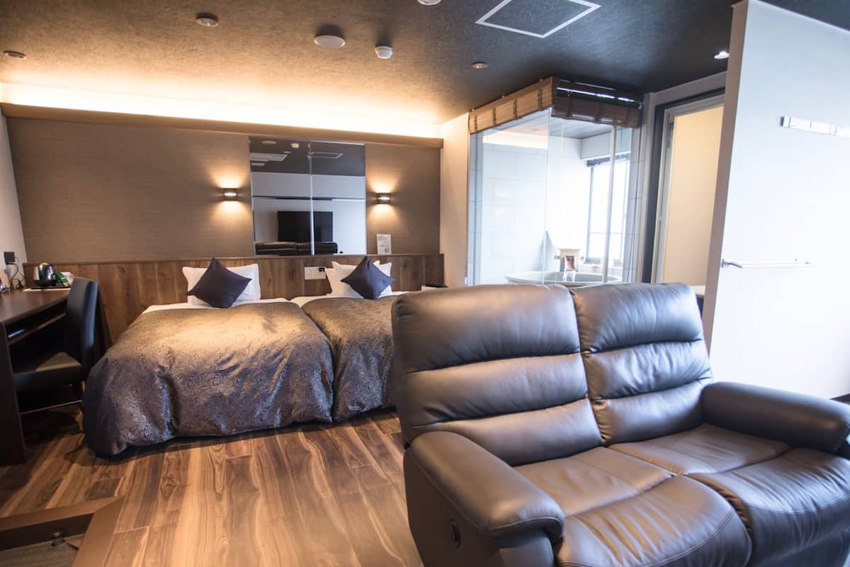 A modern hotel room with a wooden floor, a large bed with a headboard, a leather armchair, and a desk with a chair.
