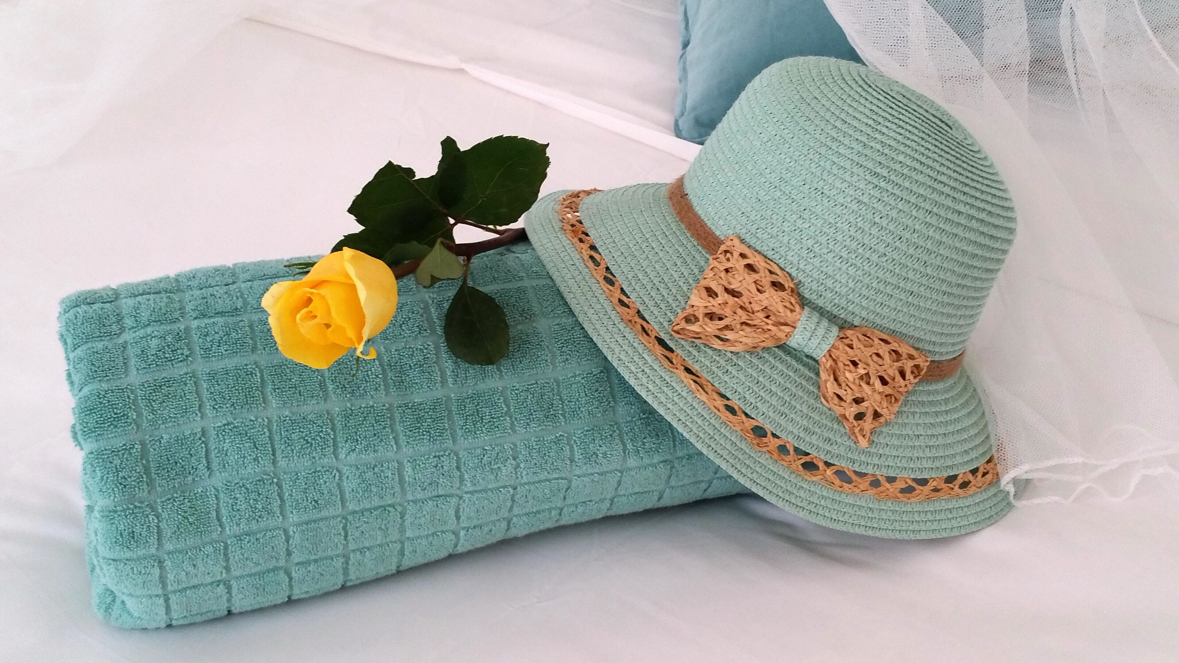 A bed with a teal textured blanket, a yellow rose, and a crocheted hat with lace details.