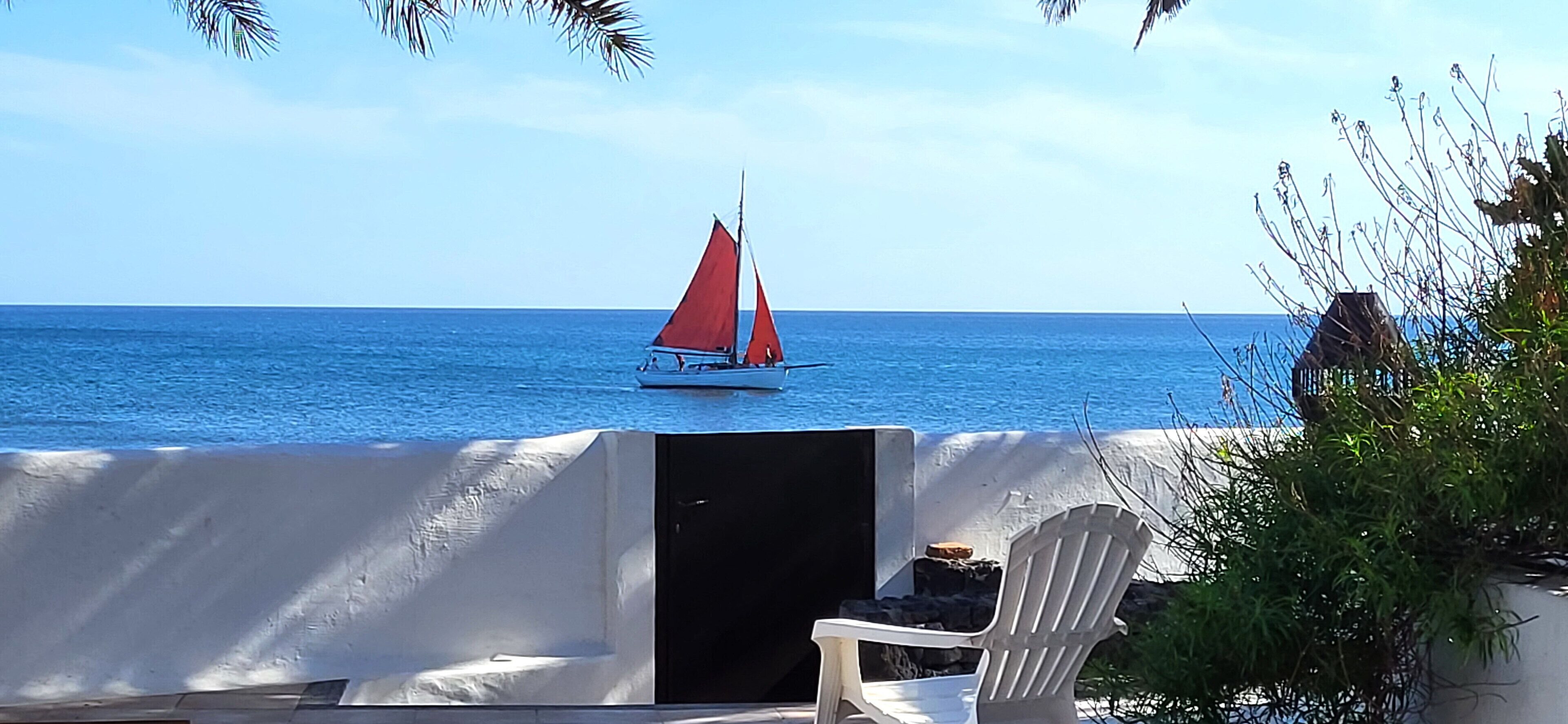Uitzicht op het strand met een zeilboot op het water, een overdekt zitgedeelte met een stoel, en een gebouw in de verte.