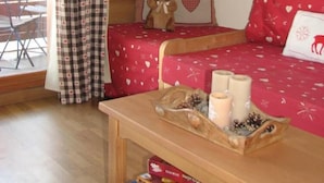Une pièce chaleureuse avec un mur en bois, un rideau à carreaux, un lit avec une literie rouge et blanche, un ours en peluche sur le lit, une petite table avec des bougies et un plateau, et une fenêtre donnant sur l’extérieur.