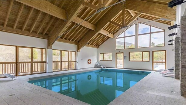 Une piscine intérieure avec des poutres en bois, de grandes fenêtres et des chaises longues.