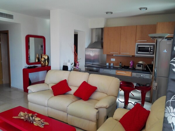 A living room with a beige sofa and red cushions, a red coffee table, and a kitchen with wooden cabinets, a microwave, and a refrigerator. - VistaMar Apartment in Estepona Málaga (Estepona)