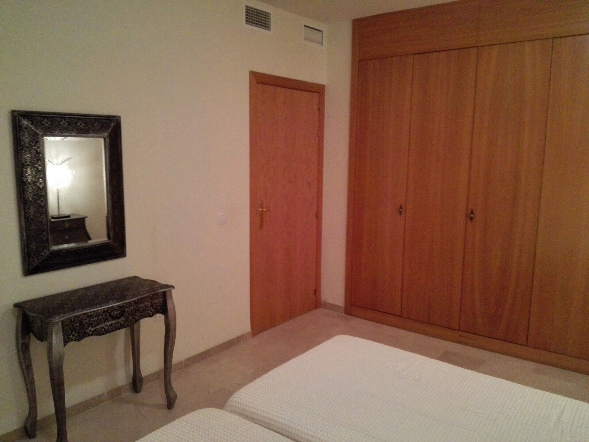 A bedroom with a bed, a wooden wardrobe, a small table, and a mirror on the wall.