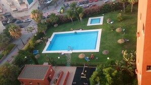 An aerial view of a residential area with a large rectangular swimming pool surrounded by a well-maintained lawn, palm trees, and a few lounge chairs. - VistaMar Apartment in Estepona Málaga (Estepona)