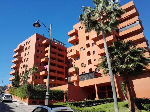 A multi-story building with balconies, palm trees, and a street with cars.