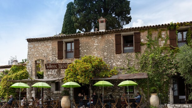 Un edificio de piedra con un letrero que dice "Le Boulangerie". Hay mesas y sillas al aire libre con sombrillas verdes, y un jarrón grande en la pared.