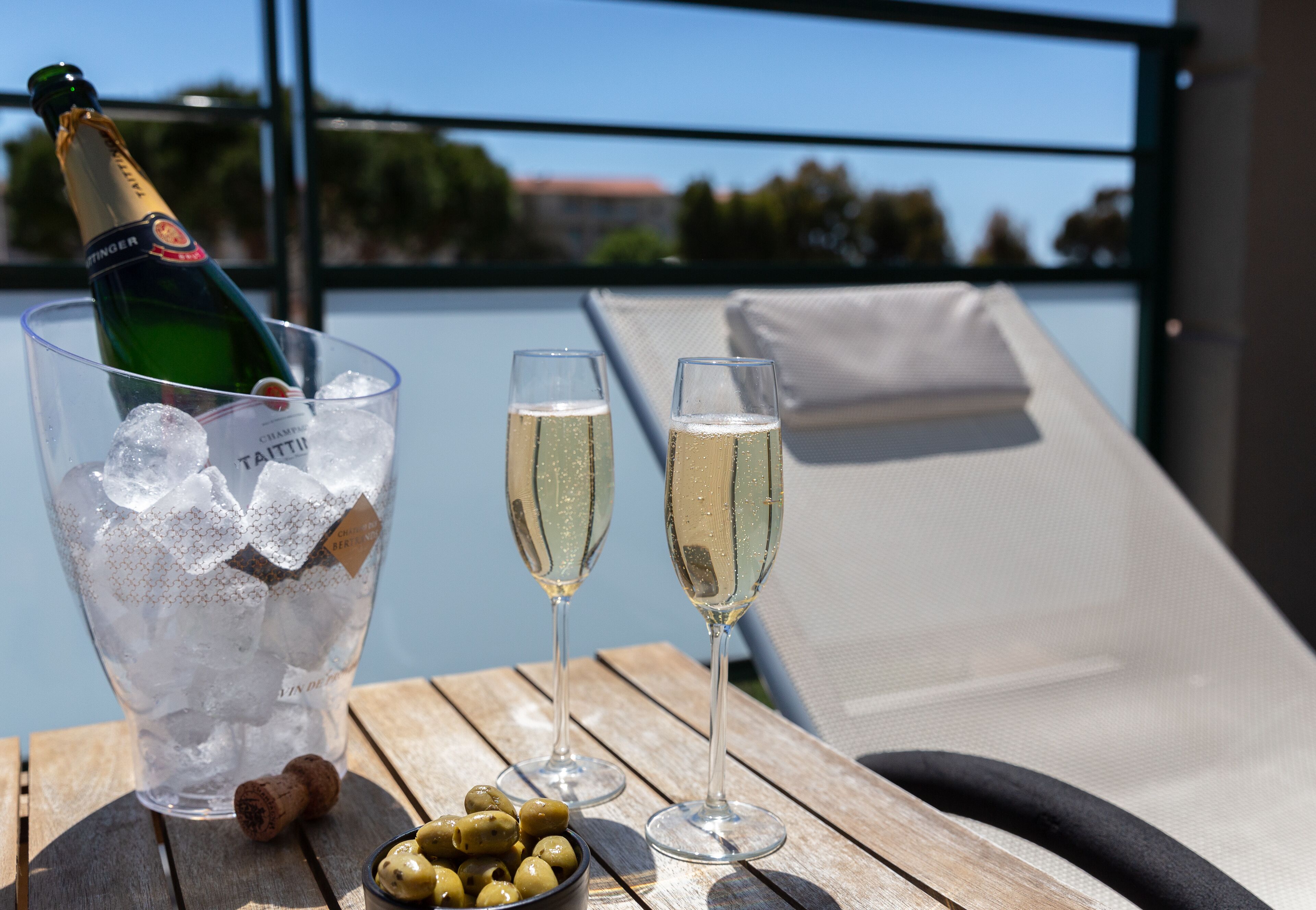 A bottle of champagne, two glasses of champagne, and a bowl of olives on a wooden table by a poolside lounge chair.