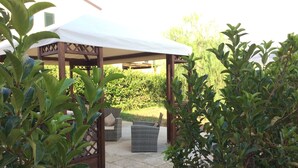 A covered outdoor seating area with a wooden pergola, stone flooring, and lush greenery.