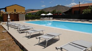 A swimming pool with sun loungers and umbrellas, surrounded by buildings and hills.