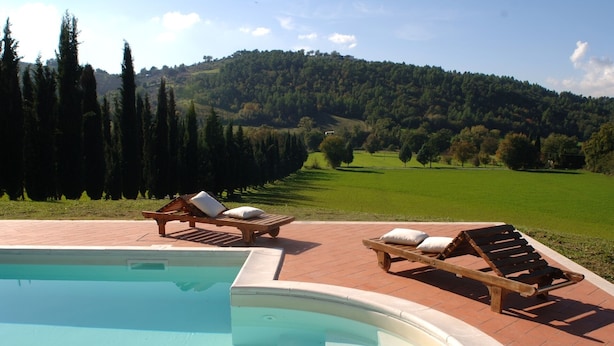 A pool area with wooden loungers and a view of a green landscape with trees and hills.