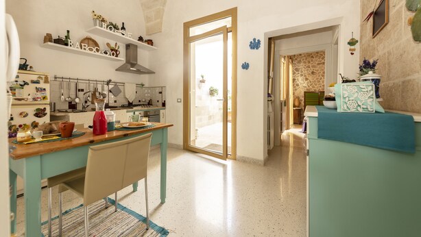 Une cuisine avec une table en bois, des chaises et divers ustensiles de cuisine. Une porte donne accès à une autre pièce.