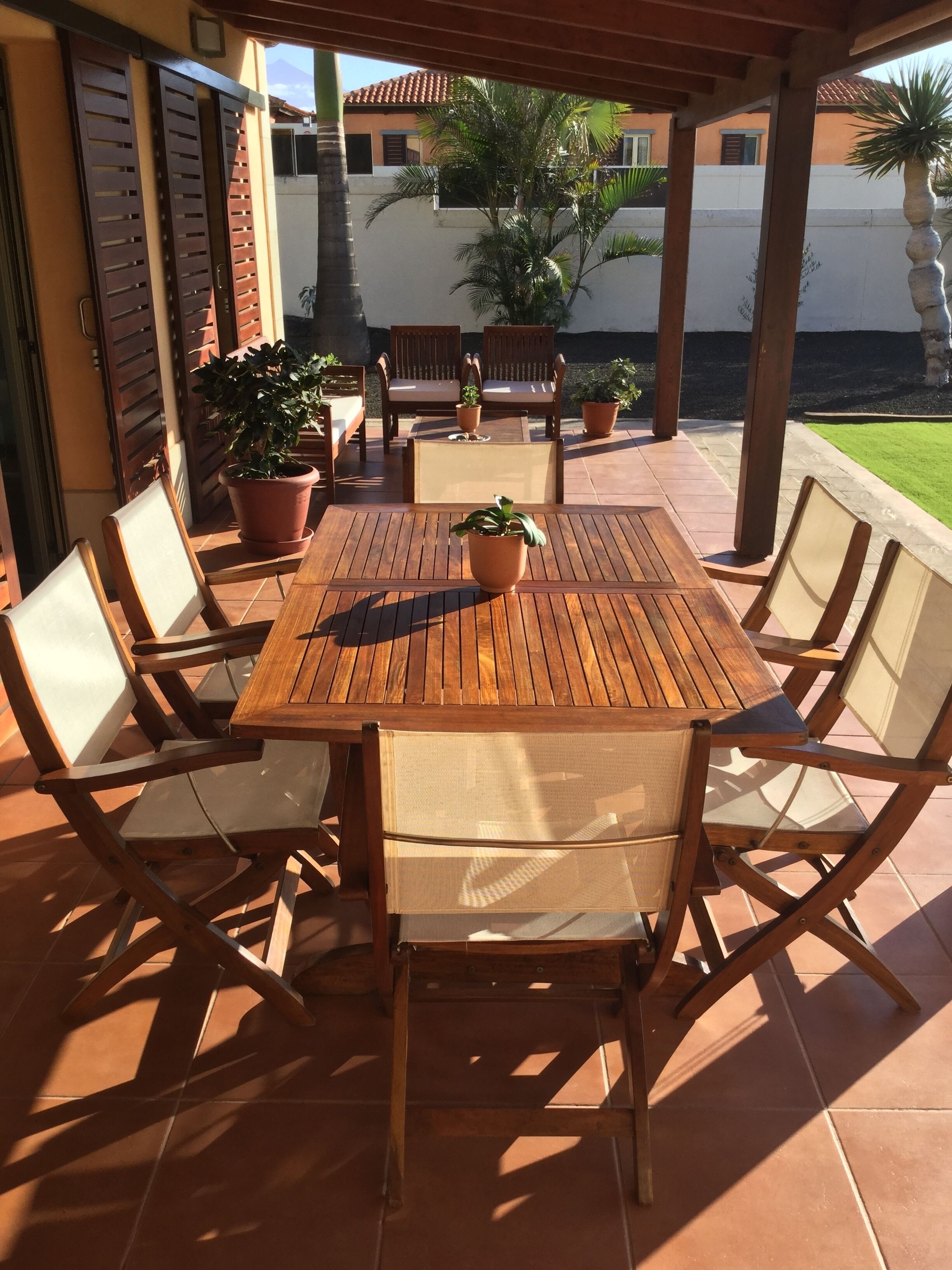 Un patio exterior con una mesa y sillas de madera, rodeado de plantas en macetas y vistas a un edificio al fondo.