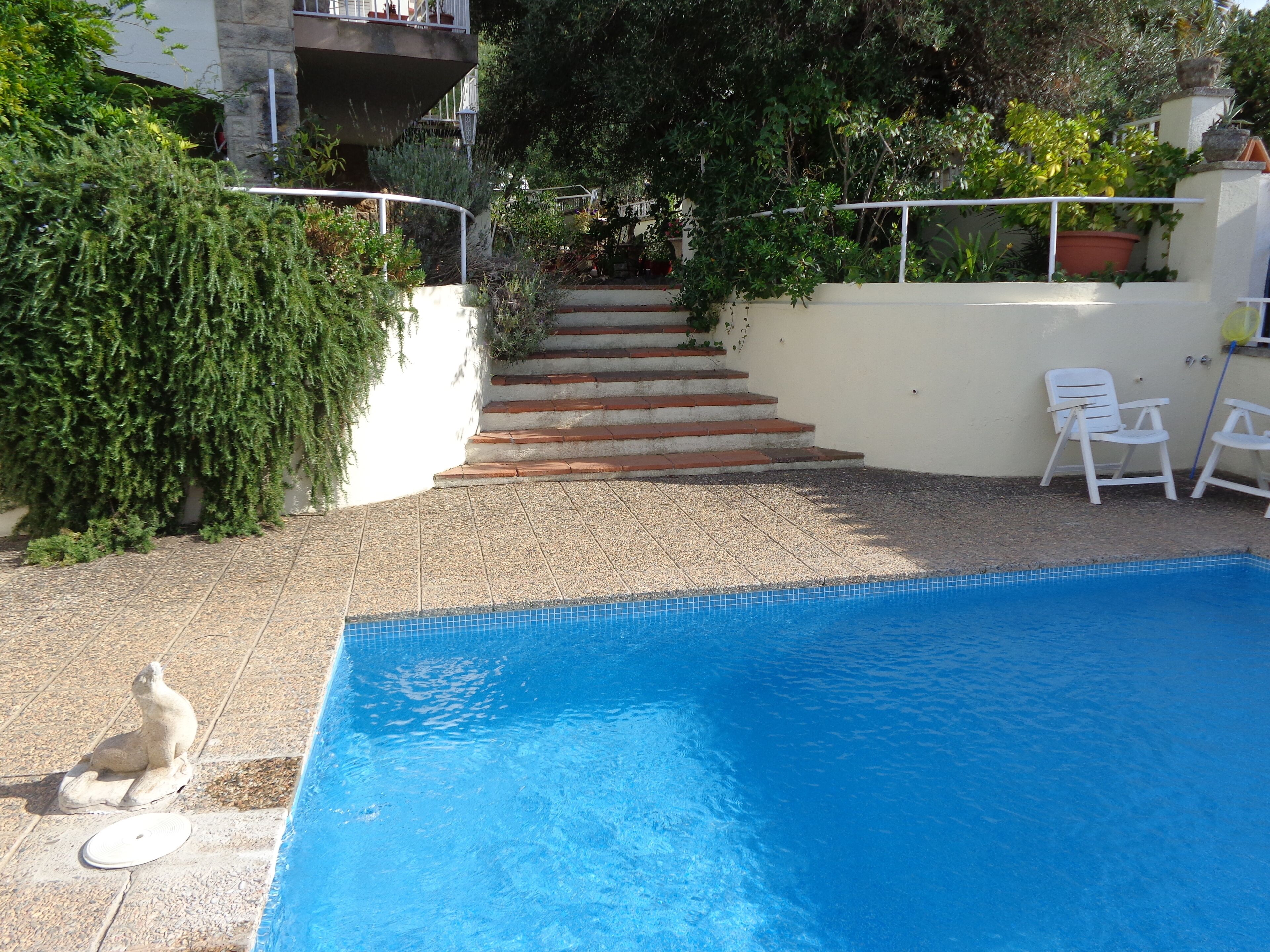 Una piscina con escalones que llevan a un balcón, rodeada de vegetación y sillas blancas.