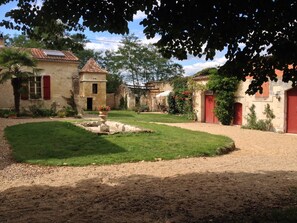 Une cour bordée de bâtiments en pierre, un chemin de gravier et une petite fontaine.