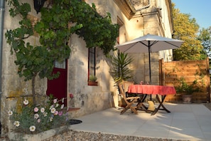 Un bâtiment en pierre avec une porte rouge, une table en bois recouverte d’une nappe rouge et un parasol blanc.