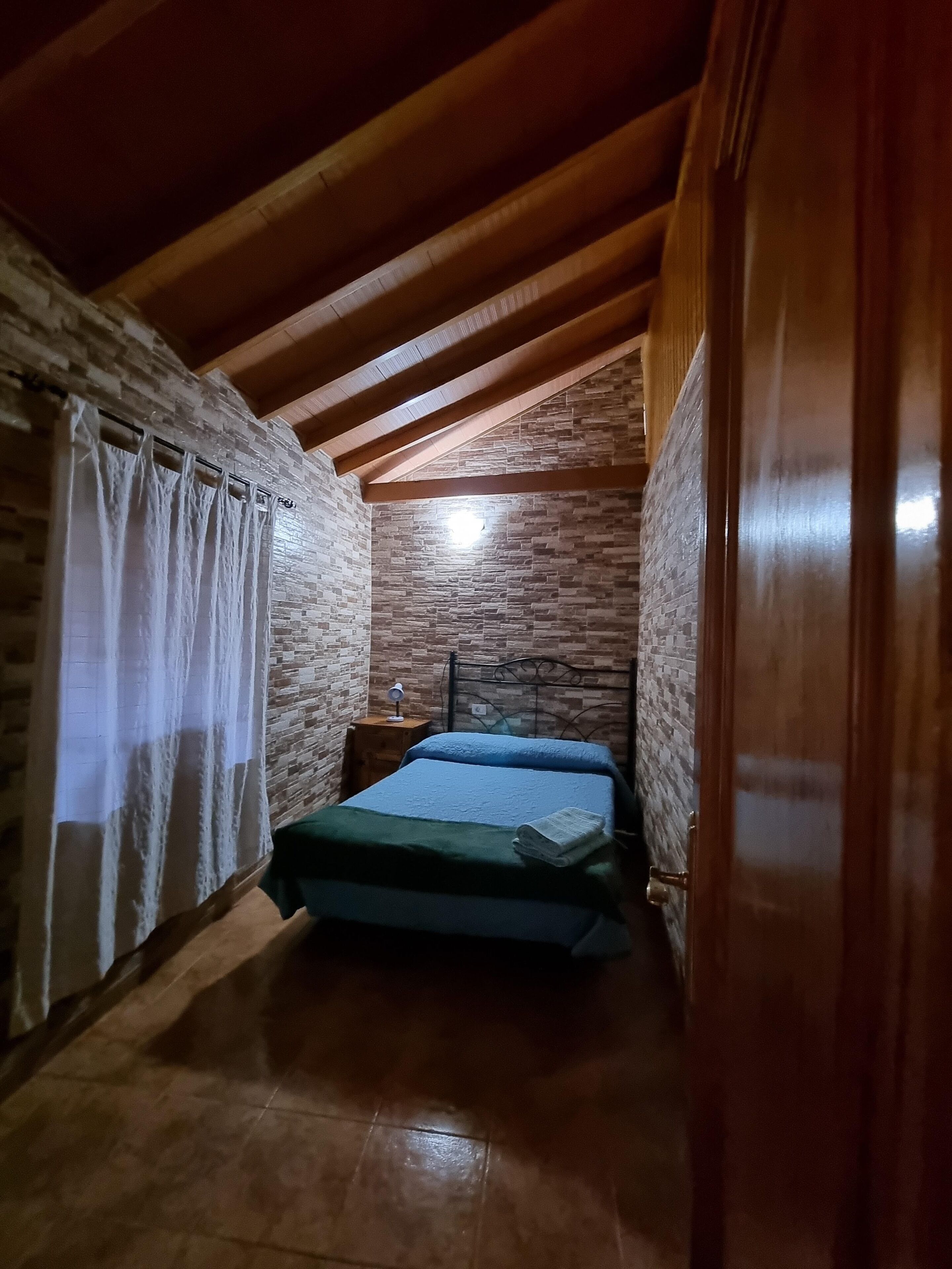 Ein kleiner, gemütlicher Raum mit schrägen Wänden, einem Bett mit blauer Matratze, einem hölzernen Nachttisch und einer Steinwand.