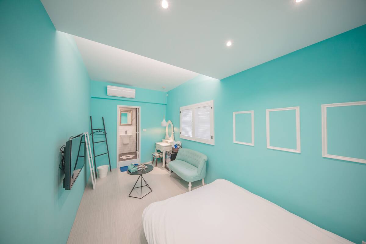 A room with teal walls, a bed, a desk, a chair, and a bathroom with a mirror and sink.