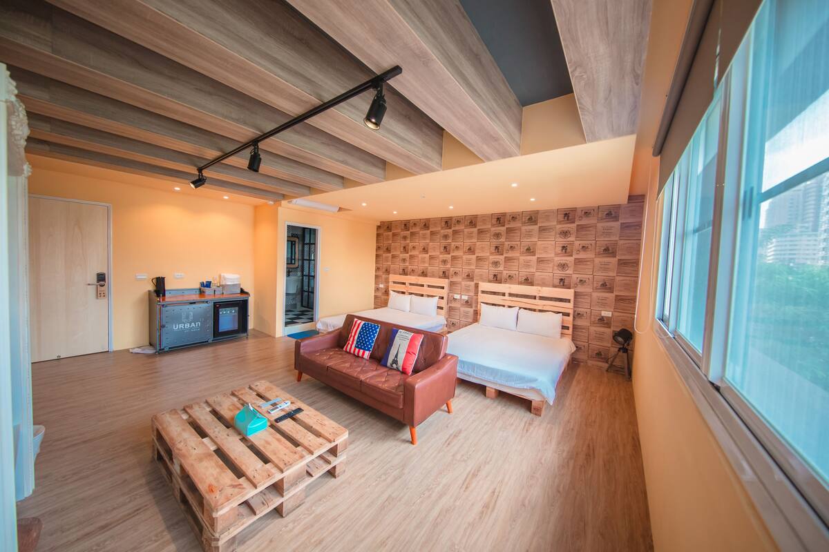 A modern hotel room with a wooden floor, a brown leather sofa, a bed with white linens, a wooden headboard, and a city view through large windows.