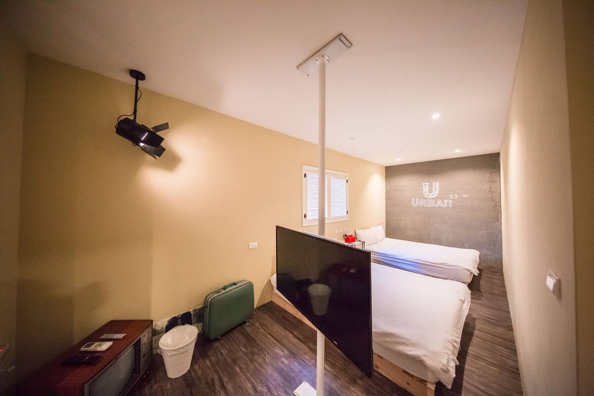 A compact hotel room with a bed, a desk with a computer, a small TV, a green suitcase, and a trash bin.