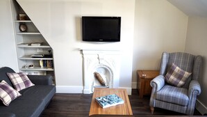 A living room with a dark wood floor, a grey sofa with plaid cushions, a plaid armchair, a wooden coffee table with a book, a fireplace, and a flat-screen TV mounted above it.