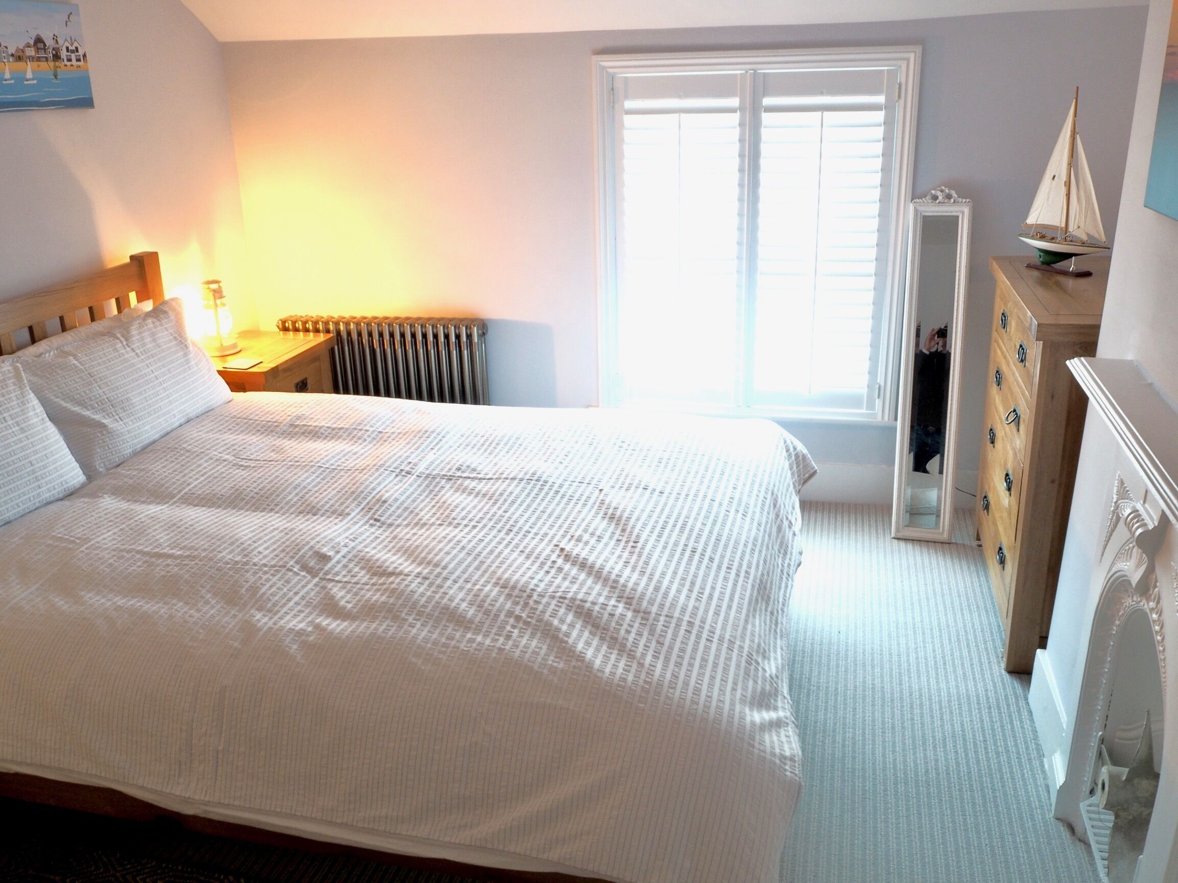 Een slaapkamer met een groot bed, een radiator, een raam met jaloezieën, een dressoir en een klein modelzeilboot erboven.