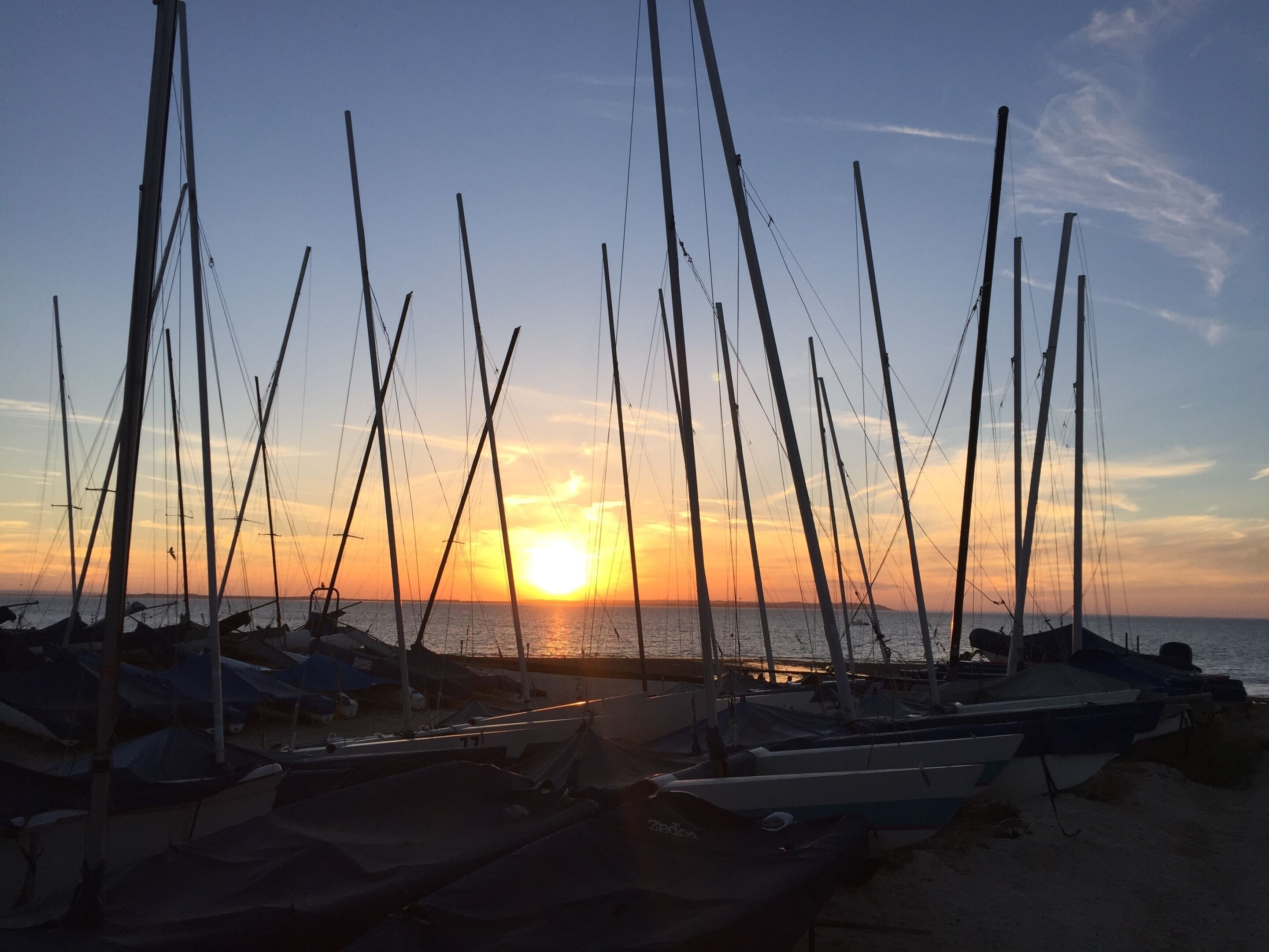 Un port de plaisance où sont amarrés de nombreux voiliers, un coucher de soleil en arrière-plan et un ciel dégagé.