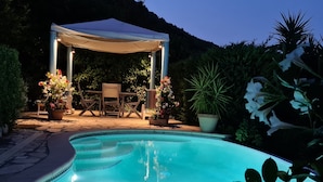 Zona de piscina con un espacio de descanso cubierto, rodeada de plantas en macetas y montañas al fondo.