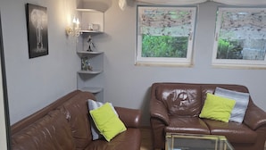 Un salon avec deux canapés en cuir marron, une table basse en verre, une lampe fixée au mur et deux fenêtres ornées de rideaux à motifs floraux.