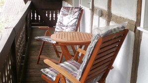 Una terraza de madera con dos sillas y una mesa de madera, un farol y una pared blanca con vigas de madera.