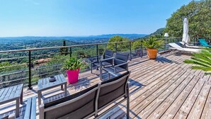 A wooden deck with outdoor seating, potted plants, and a view of distant hills. - Spacious air-conditioned villa, heated pool and SPA, breathtaking views. (Grasse)