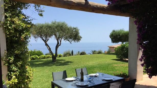Un espace de restauration en plein air avec une table dressée pour quatre personnes, offrant une vue sur un paysage verdoyant et la mer.