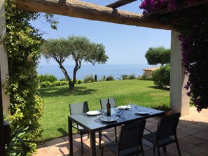 An outdoor dining area with a table set for four, overlooking a grassy landscape and the sea.