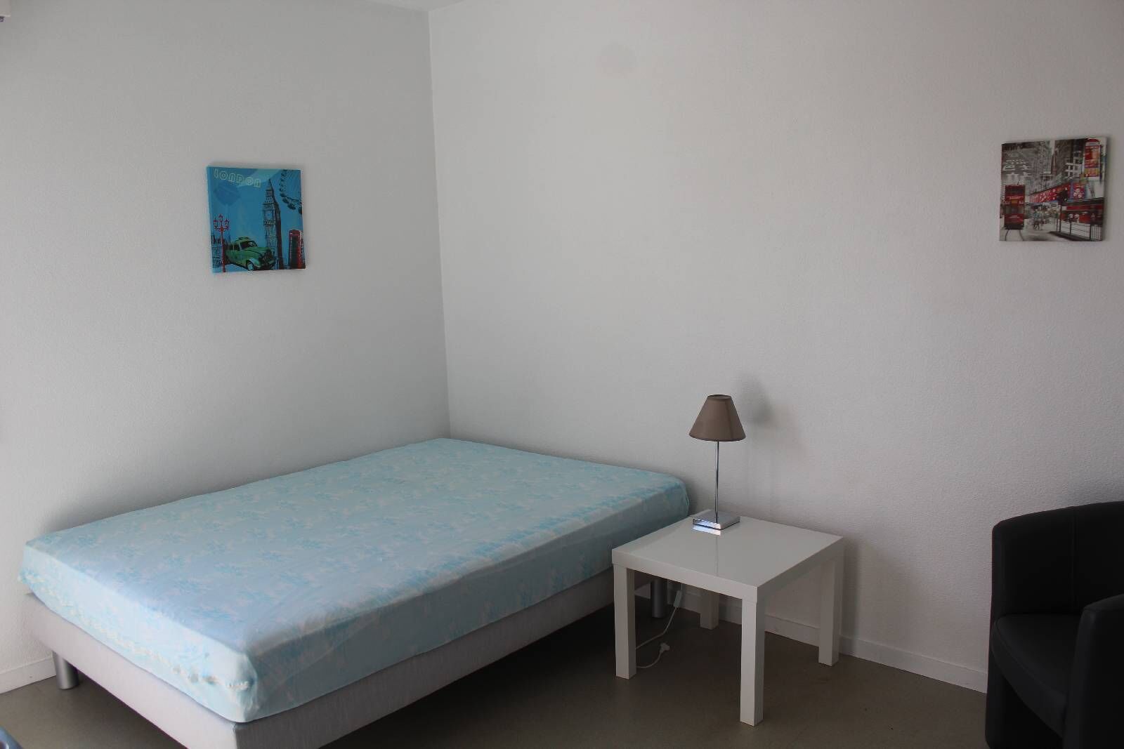 A small, single-bed room with a blue mattress, a bedside table with a lamp, and two wall-mounted pictures.