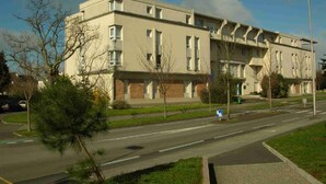Um edifício de vários andares com telhado plano e janelas retangulares, cercado por um gramado bem cuidado e algumas árvores.