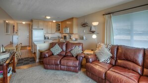 A compact living space with a kitchenette, a brown leather sofa set, a dining table with chairs, and a window with blinds.
