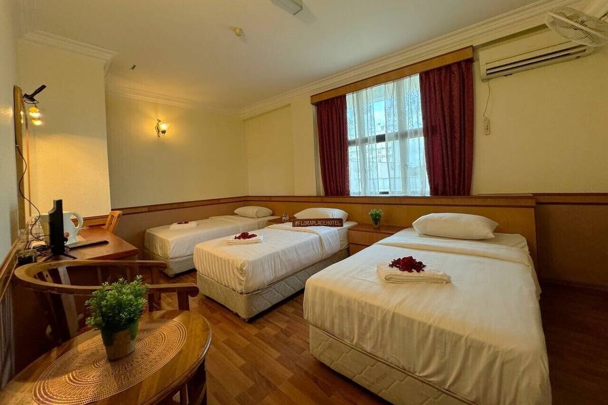 Quarto de hotel com duas camas de solteiro, uma escrivaninha com cadeira, uma mesinha com planta e uma janela com cortinas.