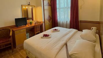 A hotel room with a bed, a desk with a computer, a chair, a mirror, and a window with curtains.