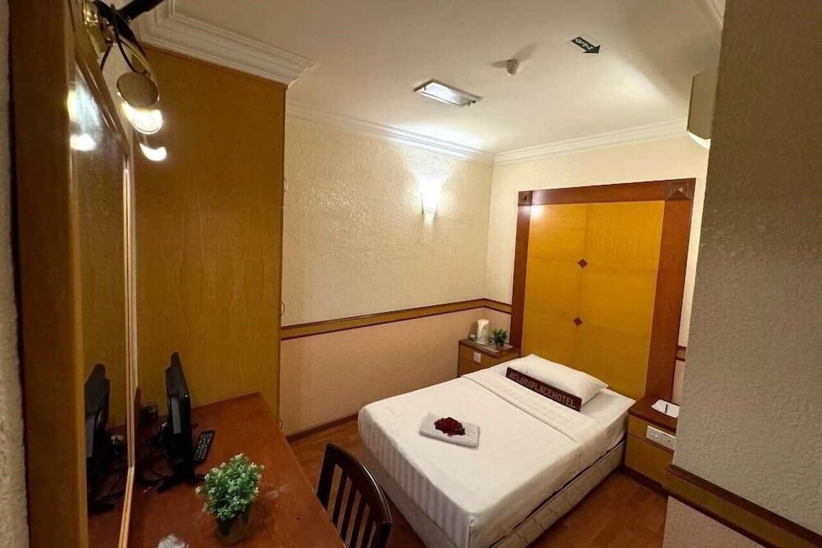 Quarto de hotel com uma cama, uma escrivaninha com um computador, uma planta pequena e uma mesa de cabeceira com abajur.