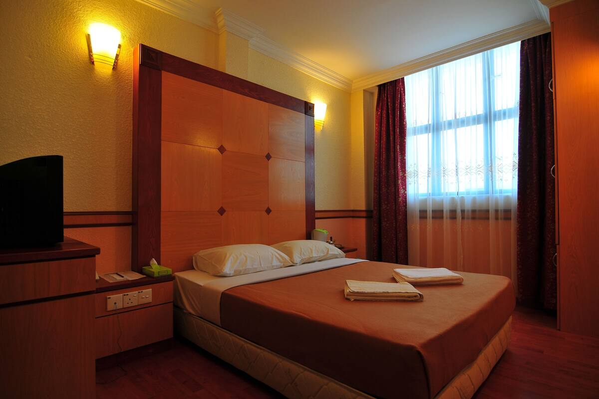 Quarto de hotel com cama, mesa de cabeceira, abajur e janela com cortinas.