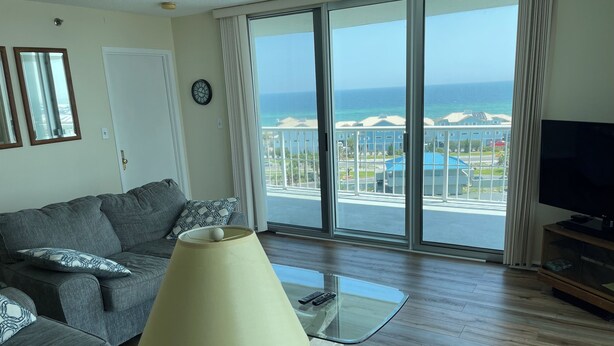 Ein Wohnzimmer mit einer Couch, einer Lampe, einem Fernseher und einem großen Fenster mit Blick auf das Meer und Gebäude.