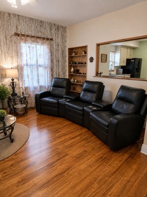 Ein Wohnzimmer mit Ecksofa, Holzboden, Esstisch mit Stühlen, einer Lampe, einem Bücherregal und einem durch einen Spiegel sichtbaren Küchenbereich.