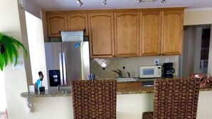 A kitchen with wooden cabinets, a stainless steel refrigerator, a microwave, and two wicker bar stools.