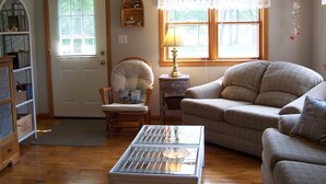 Un salon chaleureux avec un plancher en bois, un canapé beige, un fauteuil blanc, une petite table et une bibliothèque.